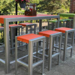 Outdoor patio with a tall metal table and eight matching stools, all with orange and green seats, surrounded by greenery and a brick wall.