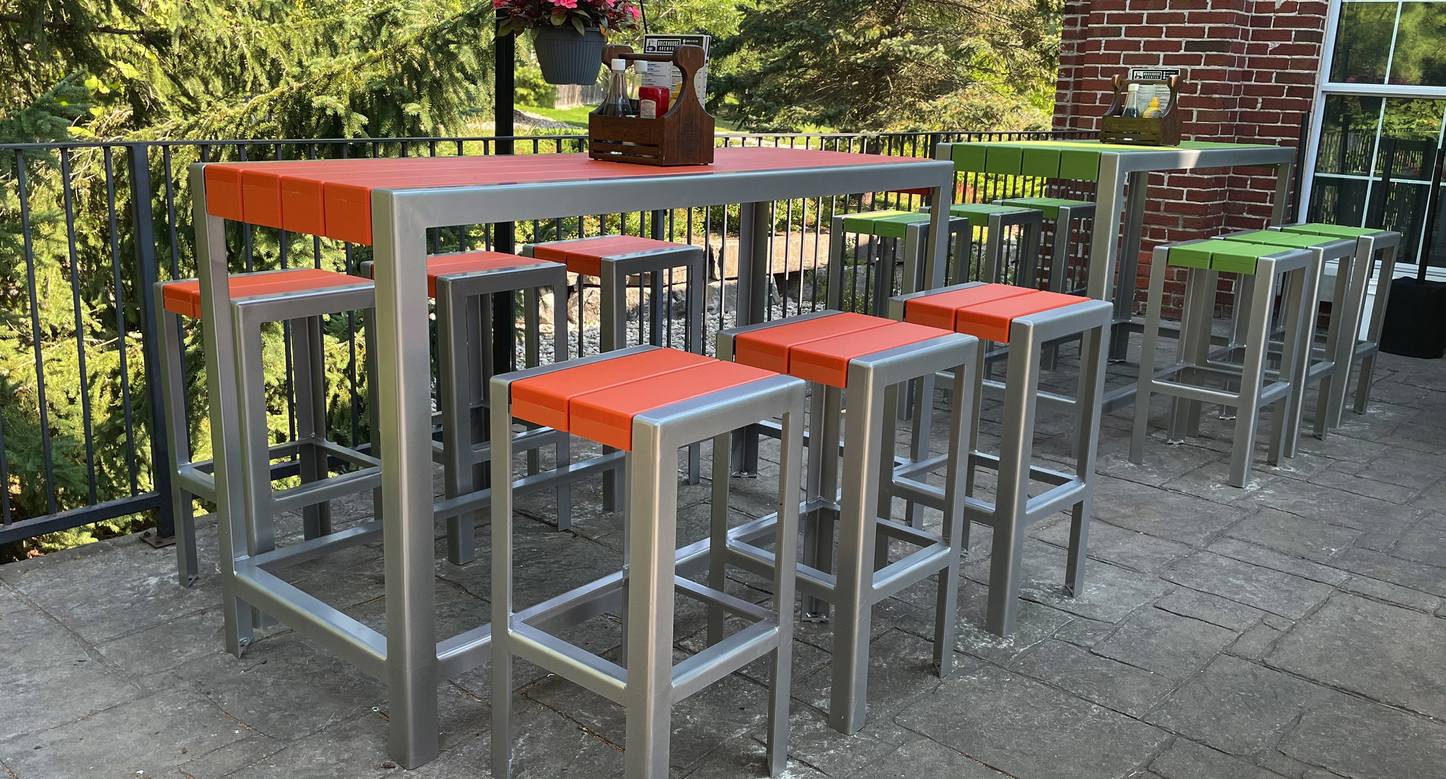 maglin_mch-1050-bar-height-stools-2 Outdoor patio with a tall metal table and eight matching stools, all with orange and green seats, surrounded by greenery and a brick wall.