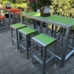 Outdoor seating area with metal-framed tables and stools, featuring green and orange slats, on a paved patio near a black metal fence and greenery.