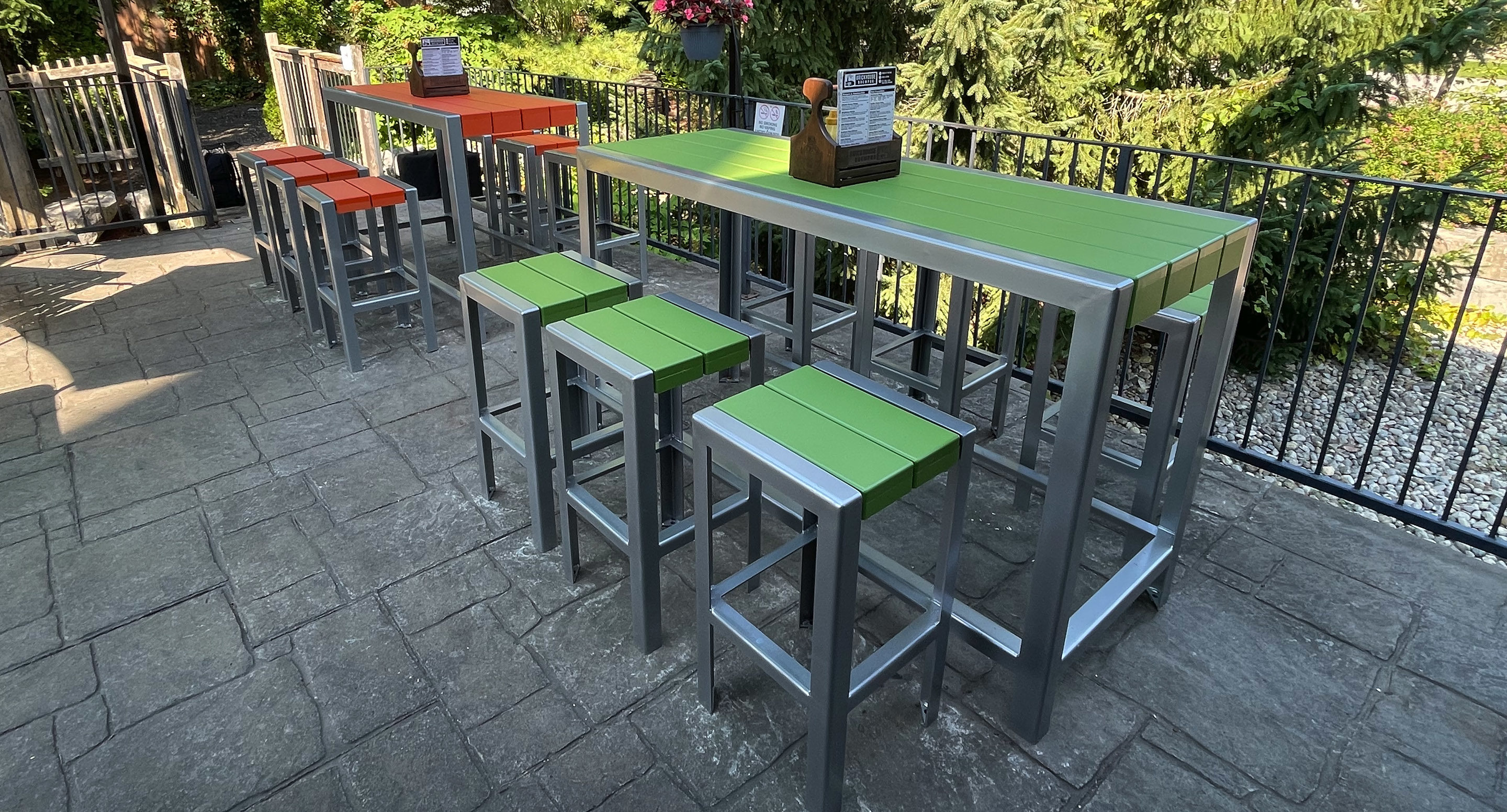 maglin_mch-1050-bar-height-stools-3 Outdoor seating area with metal-framed tables and stools, featuring green and orange slats, on a paved patio near a black metal fence and greenery.