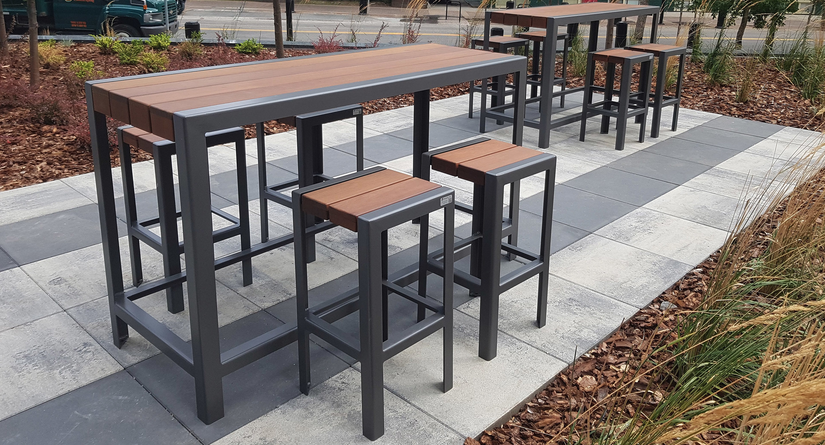 maglin_mch-1050-bar-height-stools-4 Modern outdoor seating area with metal and wood high tables and stools arranged on a tiled patio, surrounded by landscaped plants and mulch.