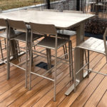 A metal outdoor dining table with six matching high chairs on a wooden deck, set in an open area with grass and fencing in the background.