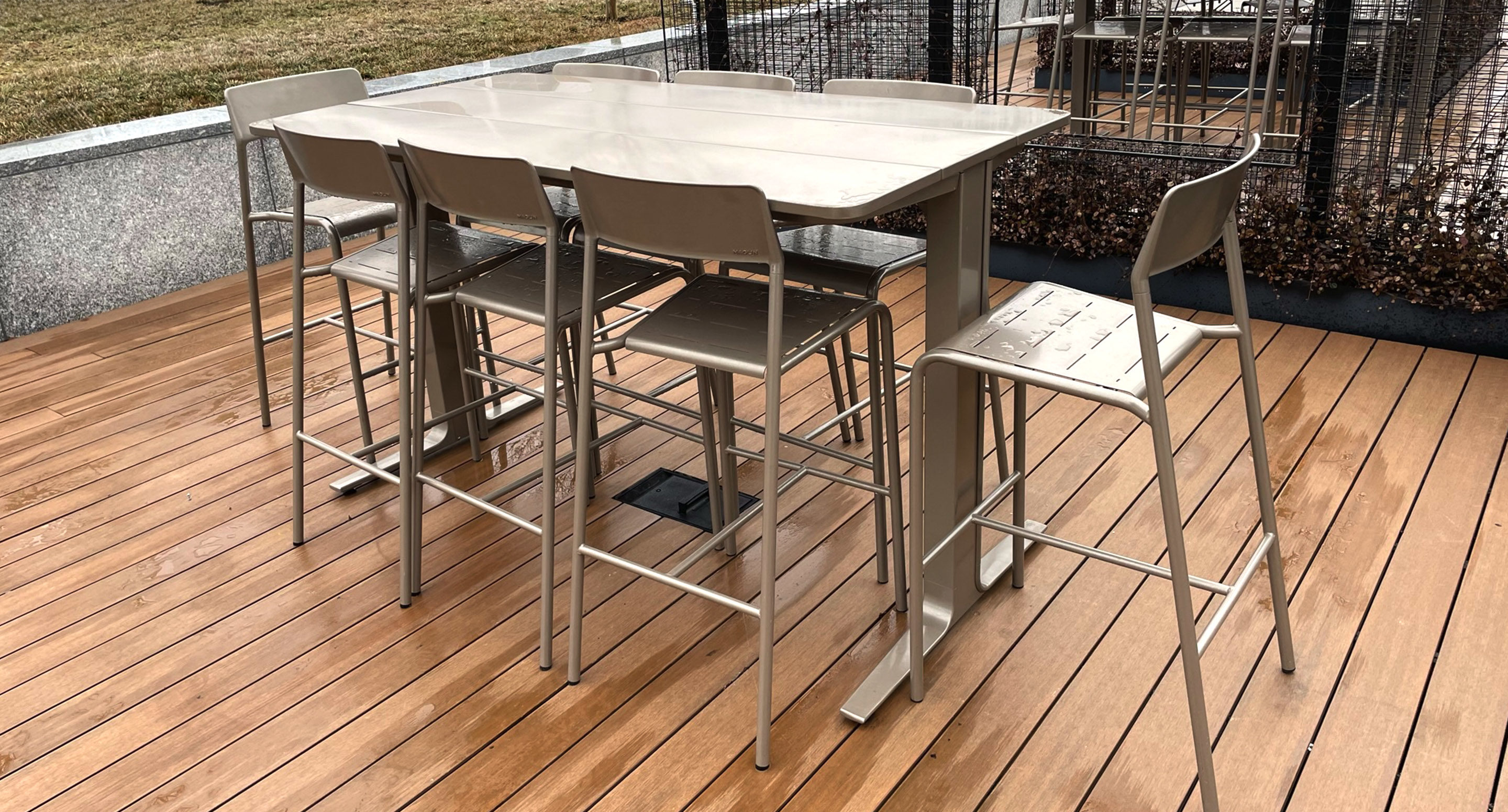 maglin_mch-1700-foro-bar-height-stool-1 A metal outdoor dining table with six matching high chairs on a wooden deck, set in an open area with grass and fencing in the background.
