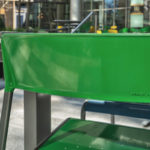 A close-up of a green metal chair at an outdoor table in a modern urban setting, with additional tables and chairs in the background.