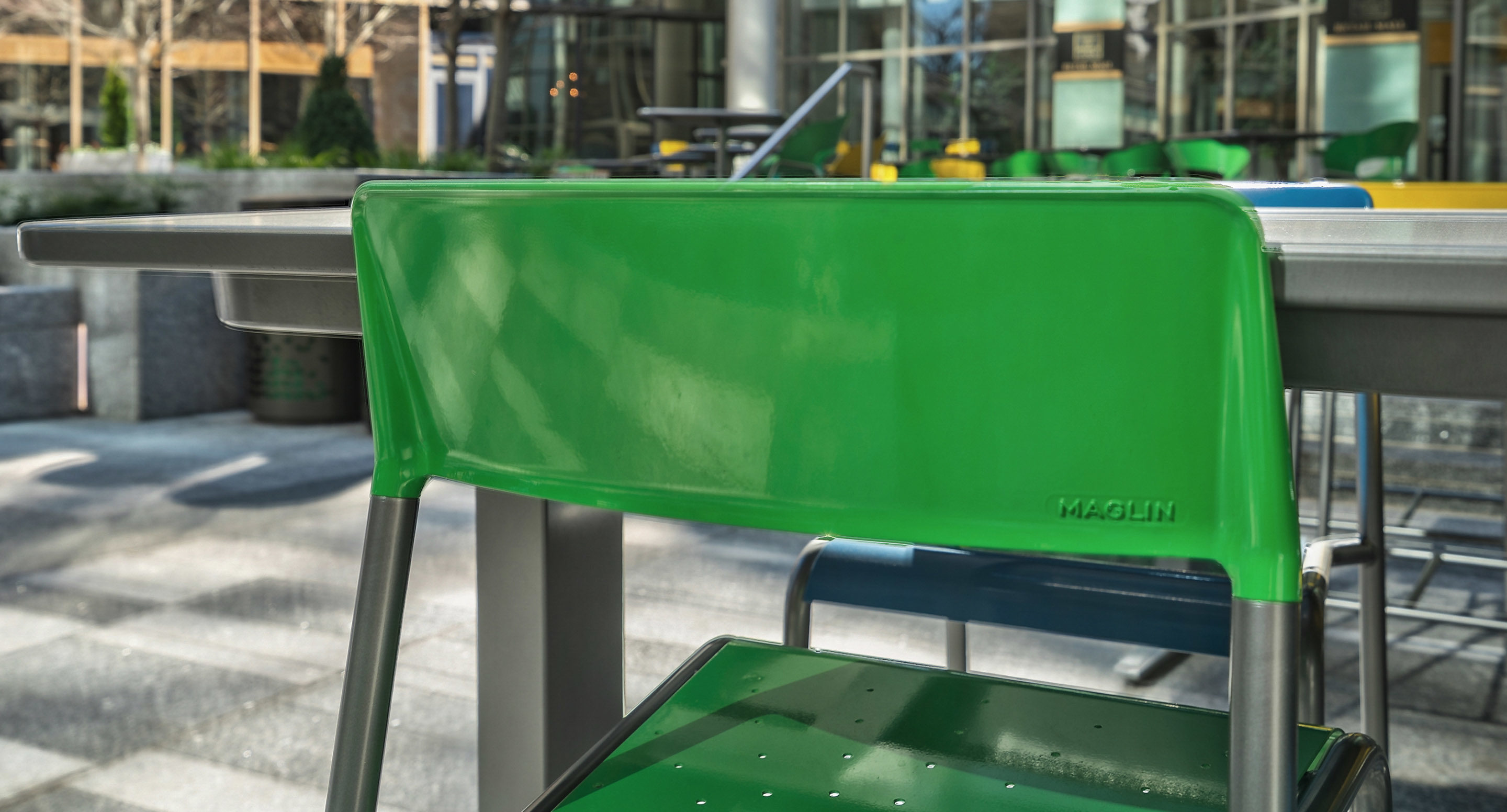 maglin_mch-1700-foro-bar-height-stool-2 A close-up of a green metal chair at an outdoor table in a modern urban setting, with additional tables and chairs in the background.