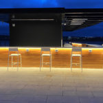 A rooftop bar with wooden paneling and white bar stools, illuminated by warm lighting, overlooks a cityscape at dusk.