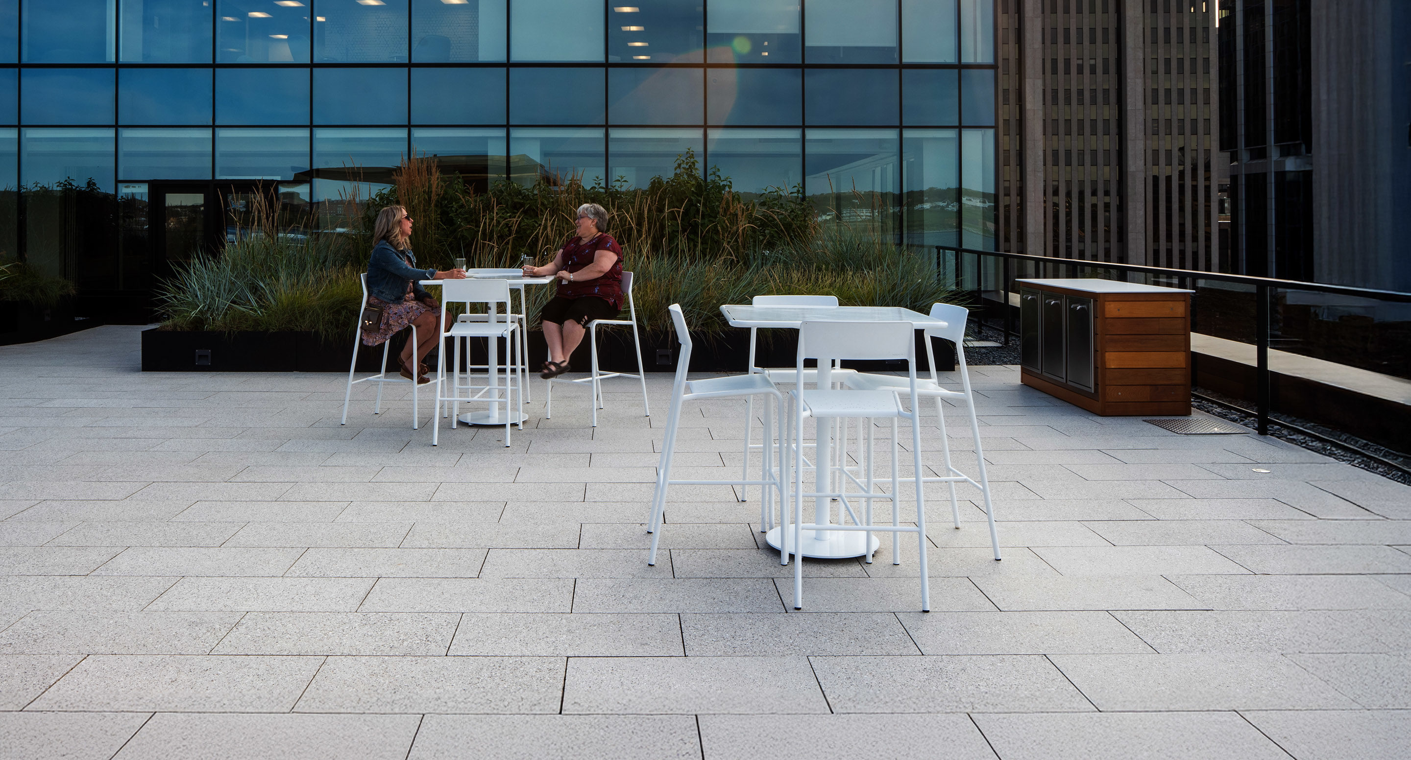 maglin_mch-1700-foro-bar-height-stool-4 Two people sit and talk at a white table on a rooftop patio with modern outdoor furniture and tall glass building windows in the background.