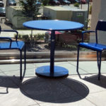 Two blue metal chairs and a round blue table are positioned indoors near a large window, casting clear shadows on the tiled floor.