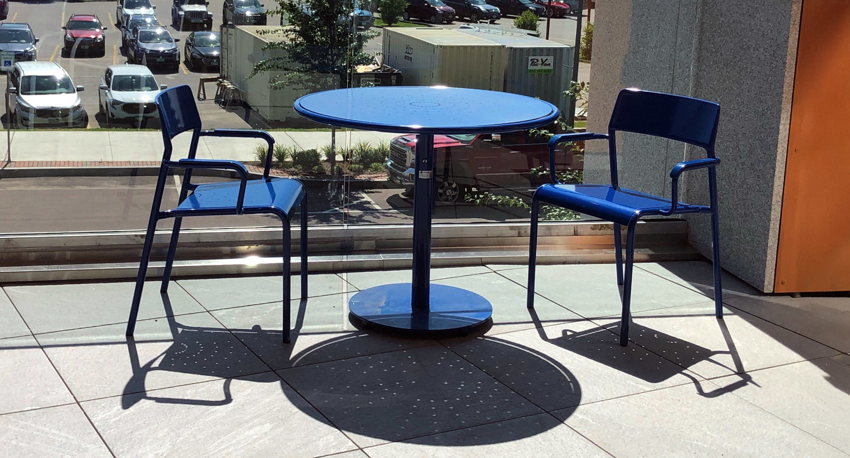 maglin_mch-1700-foro-chairs-1 Two blue metal chairs and a round blue table are positioned indoors near a large window, casting clear shadows on the tiled floor.