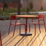 Outdoor seating area with red metal chairs and round tables on a wooden deck, lit by warm sunlight.