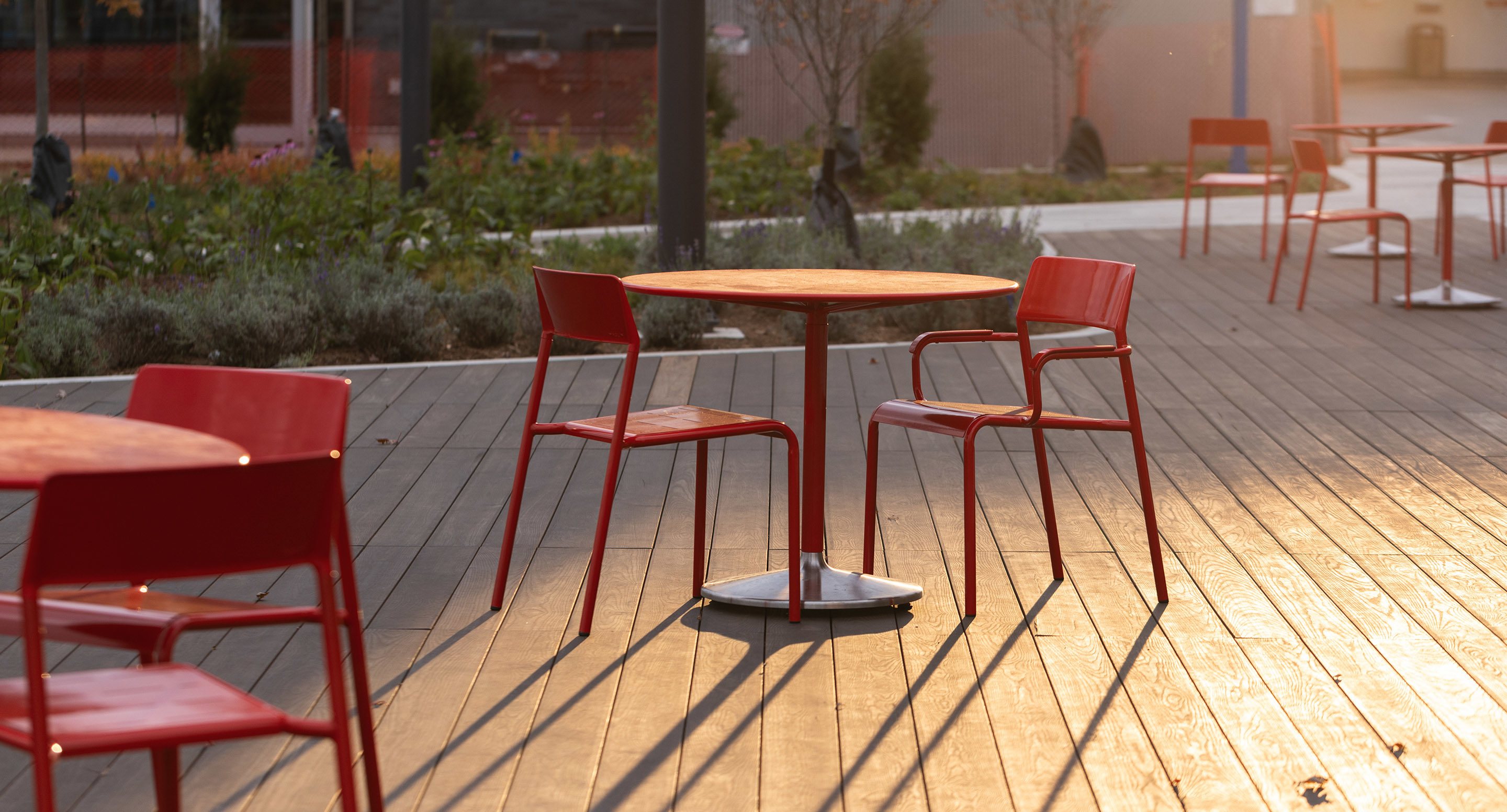 maglin_mch-1700-foro-chairs-2 Outdoor seating area with red metal chairs and round tables on a wooden deck, lit by warm sunlight.