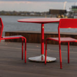 Two red metal chairs and a round red table sit on a wooden deck by the water, with buildings and a boat visible in the background.