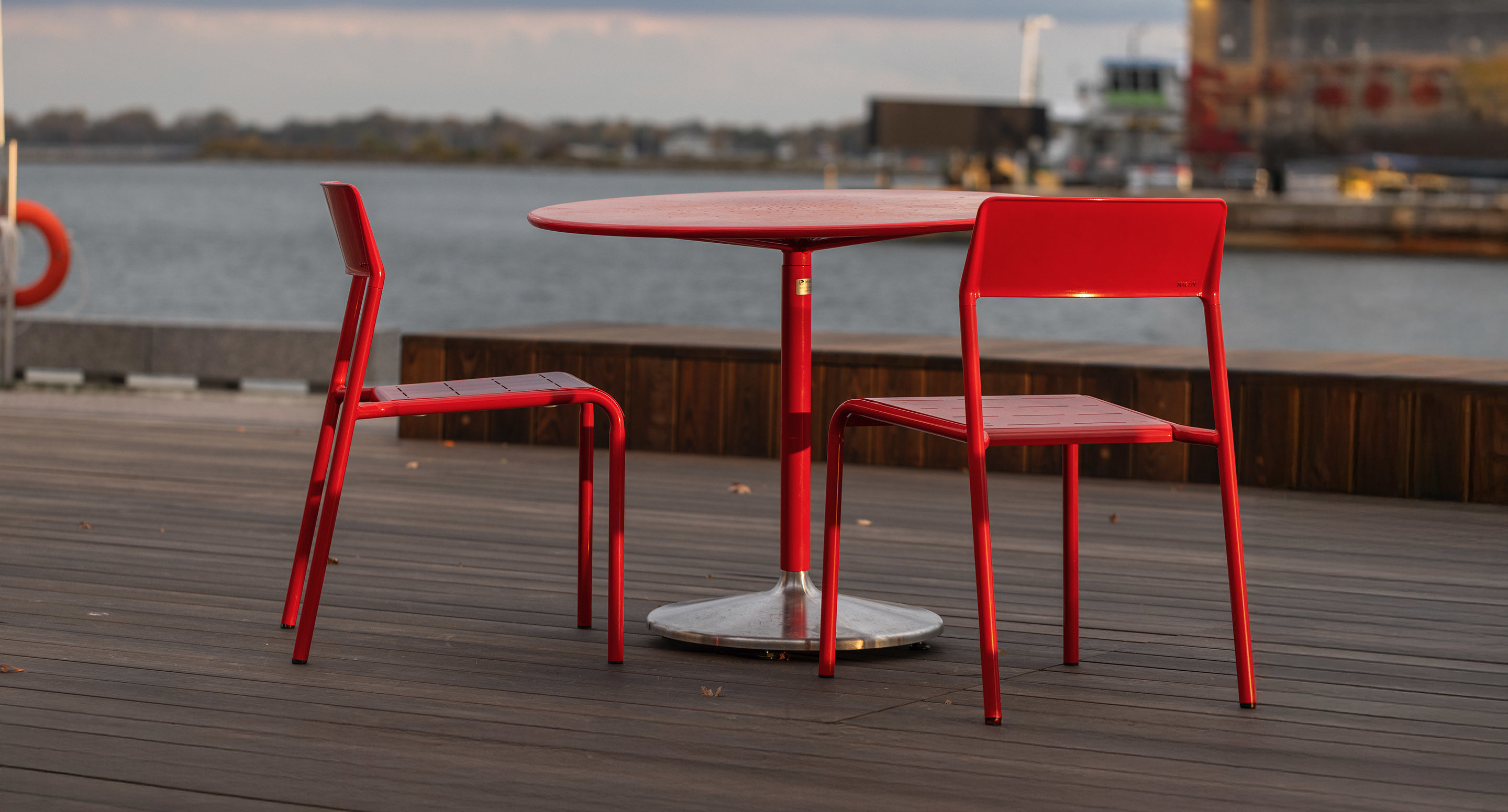maglin_mch-1700-foro-chairs-3 Two red metal chairs and a round red table sit on a wooden deck by the water, with buildings and a boat visible in the background.
