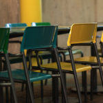 A row of green and yellow chairs is arranged around a table in an outdoor setting with a concrete wall in the background.