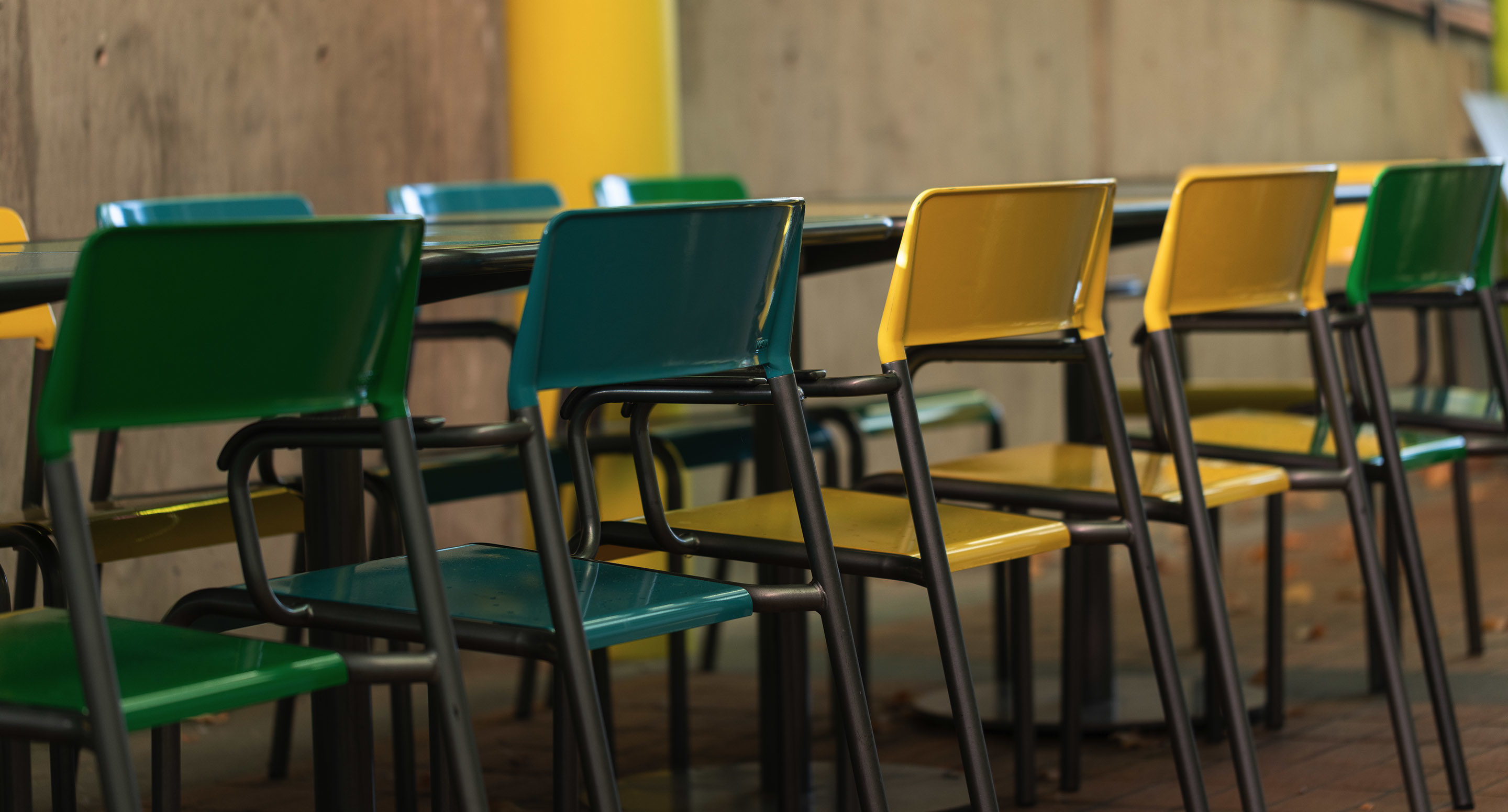 maglin_mch-1700-foro-chairs-4 A row of green and yellow chairs is arranged around a table in an outdoor setting with a concrete wall in the background.