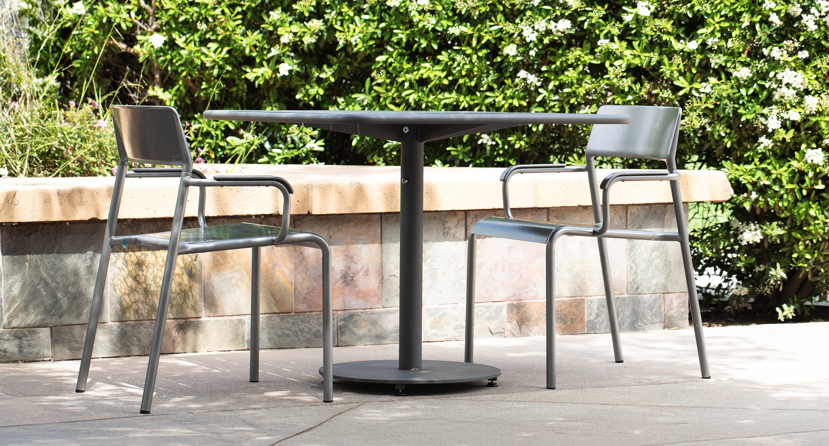 maglin_mch-1700-foro-chairs-5 Two metal chairs are placed at a round black table on a stone patio, with a hedge and flowering plants in the background.