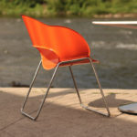 An orange plastic chair with metal legs sits beside a round white table on a concrete surface near a river.