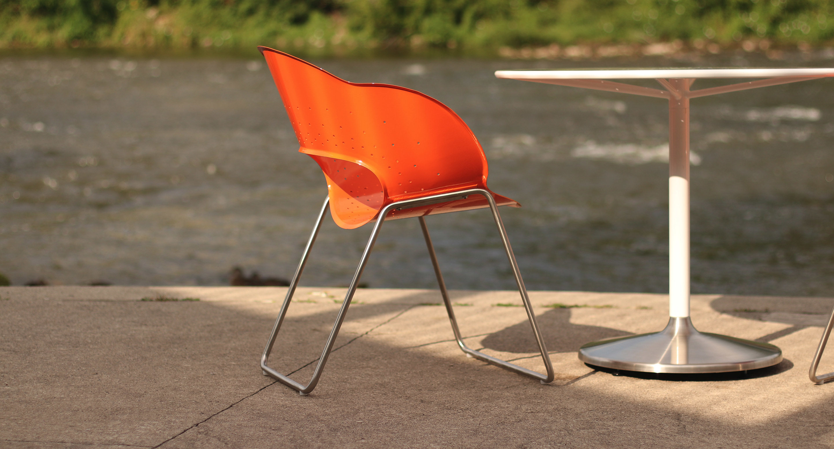 An orange plastic chair with metal legs sits beside a round white table on a concrete surface near a river.