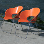 Two modern orange plastic chairs with metal legs are positioned side by side on a concrete surface near a body of water.