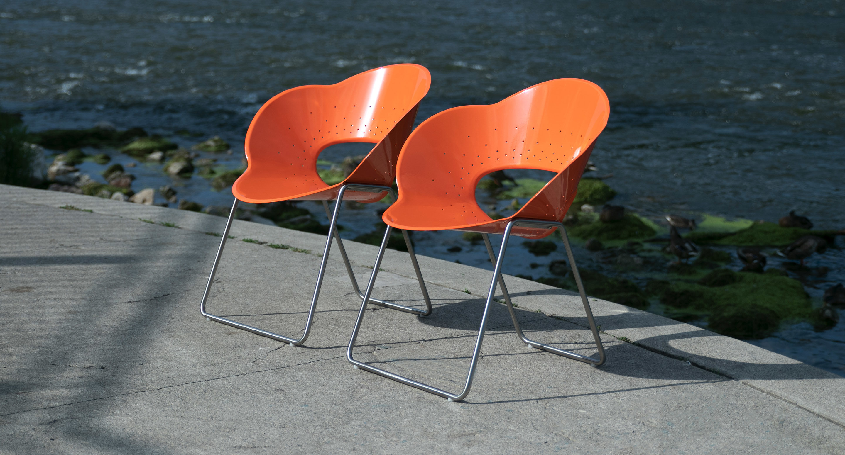 Two modern orange plastic chairs with metal legs are positioned side by side on a concrete surface near a body of water.
