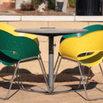 A round metal table with two green and two yellow chairs sits on a paved outdoor area near a low concrete wall.