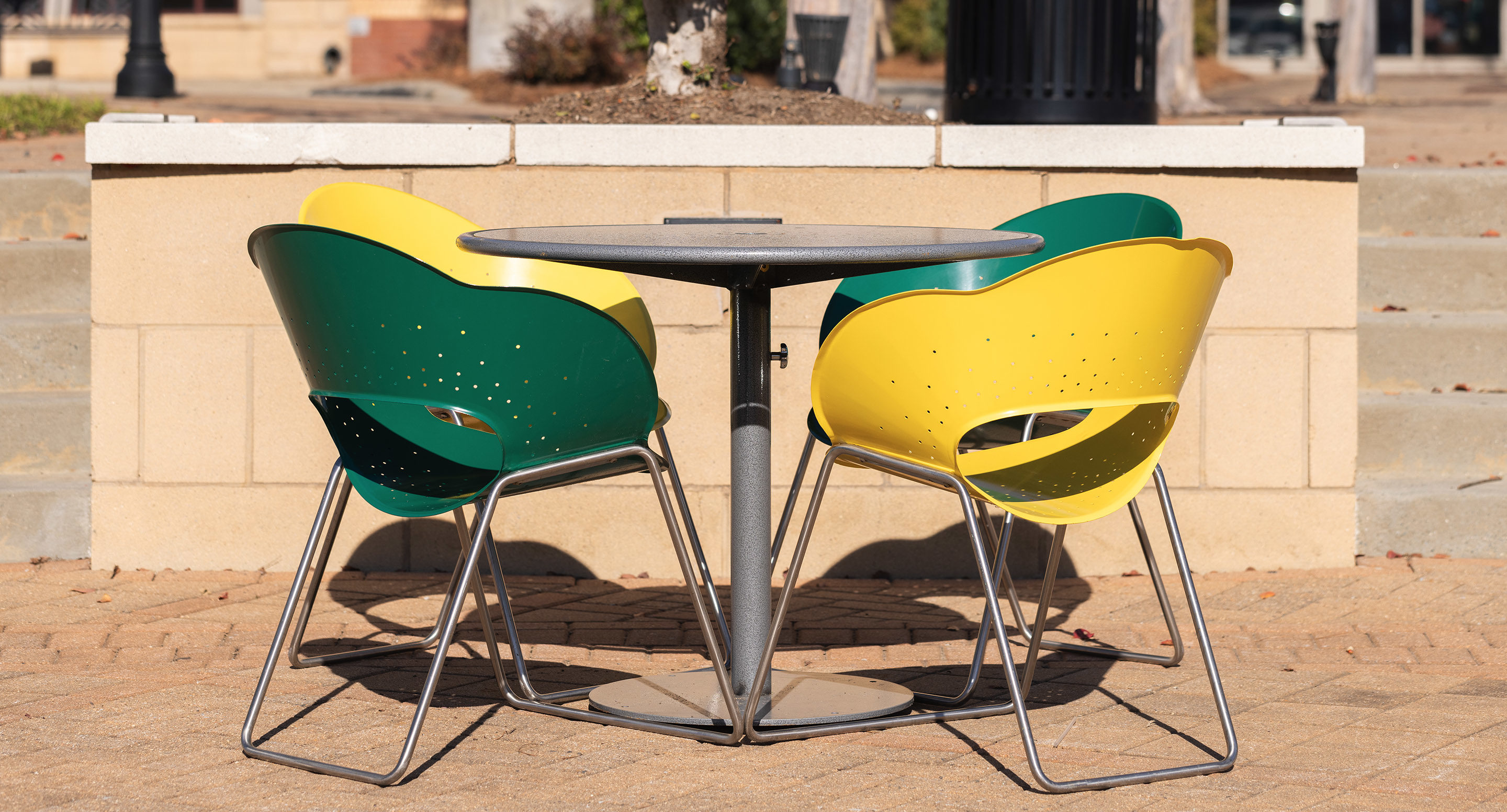 A round metal table with two green and two yellow chairs sits on a paved outdoor area near a low concrete wall.