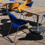 Outdoor seating area with yellow and blue plastic chairs around wooden tables on a sunlit patio; a person’s legs are visible at the left edge.