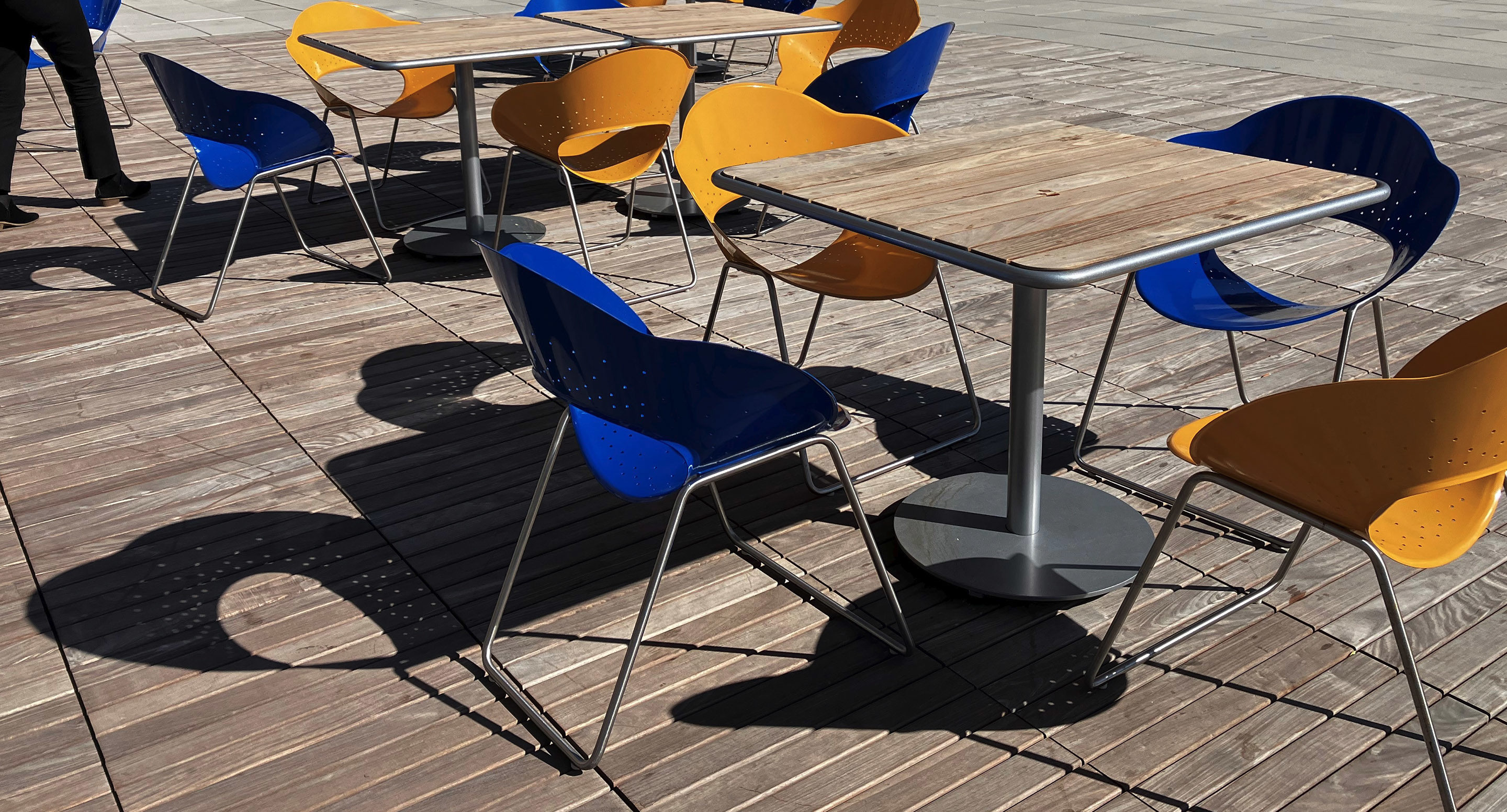 maglin_mch-1800-battery-chairs-5 Outdoor seating area with yellow and blue plastic chairs around wooden tables on a sunlit patio; a person’s legs are visible at the left edge.