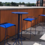 A small outdoor patio with a tall blue metal table and two matching blue metal stools next to a wooden railing.