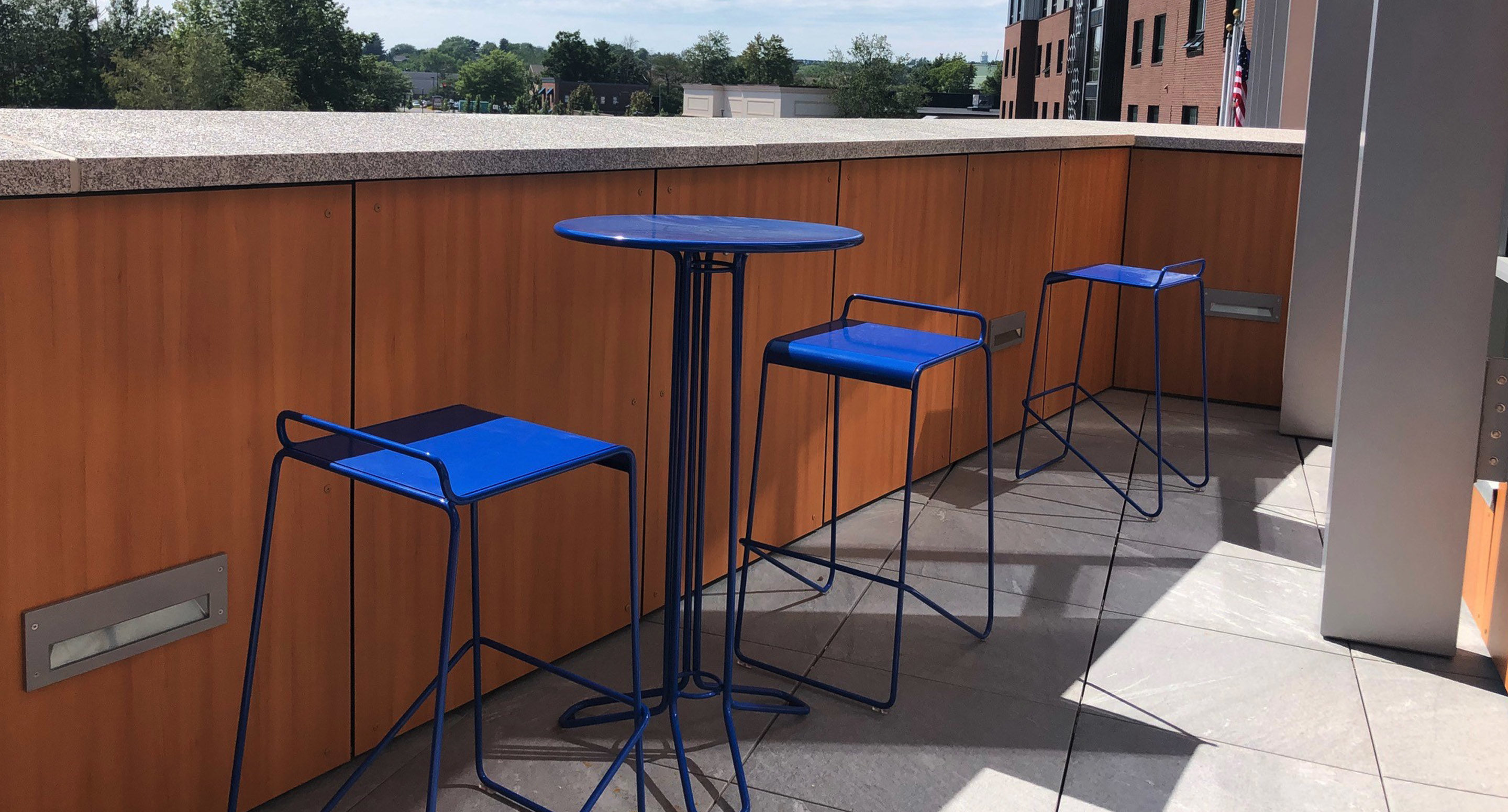 maglin_mch-2000-kontur-bar-height-stool-1 A small outdoor patio with a tall blue metal table and two matching blue metal stools next to a wooden railing.