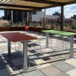 An outdoor rooftop patio features two tables, one red and one green, with stools underneath, surrounded by city buildings under a pergola structure.