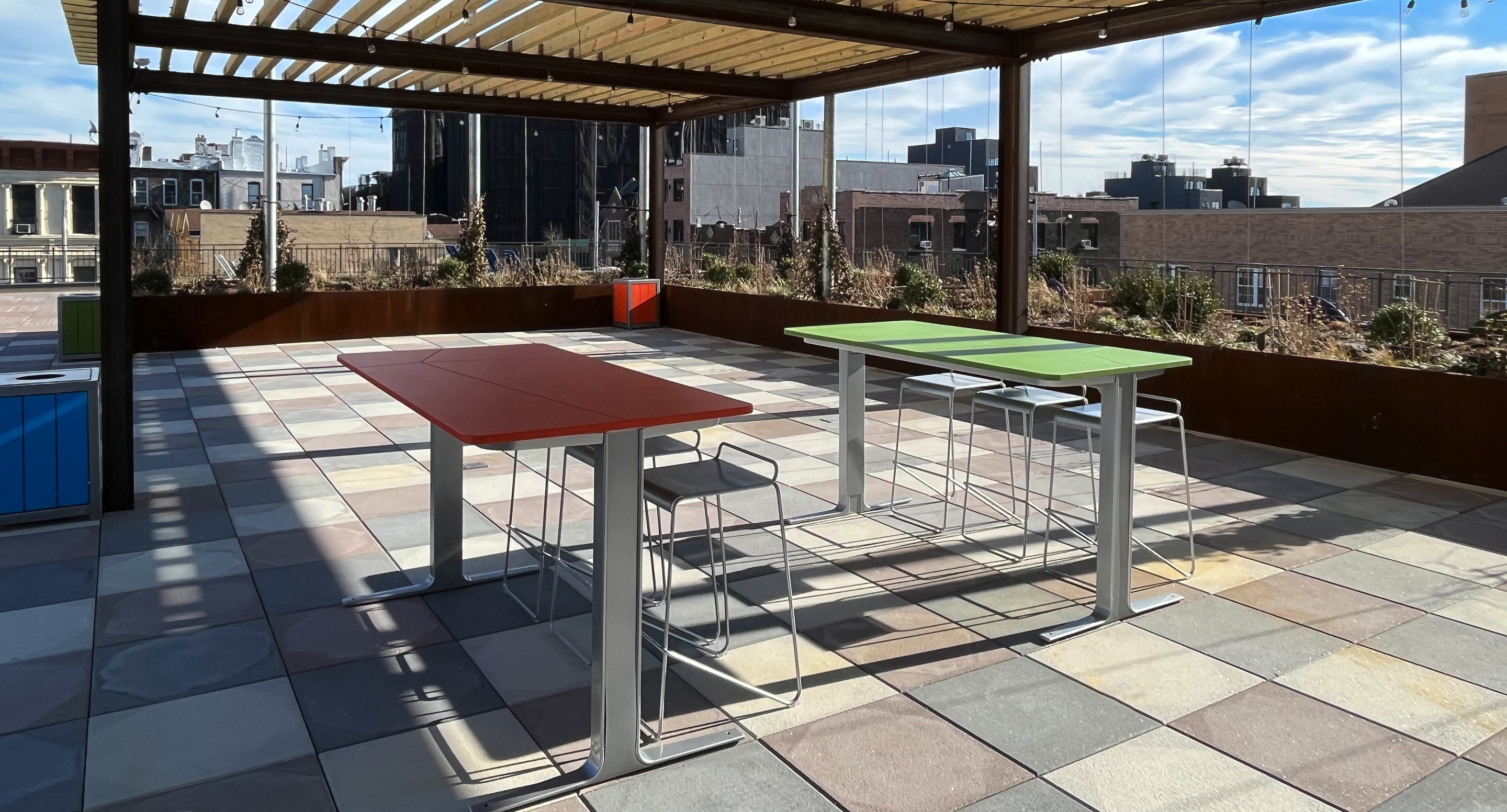 maglin_mch-2000-kontur-bar-height-stool-2 An outdoor rooftop patio features two tables, one red and one green, with stools underneath, surrounded by city buildings under a pergola structure.