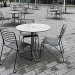 Empty metal tables and chairs are arranged on a paved outdoor plaza, with no people present and droplets of water visible on the furniture.