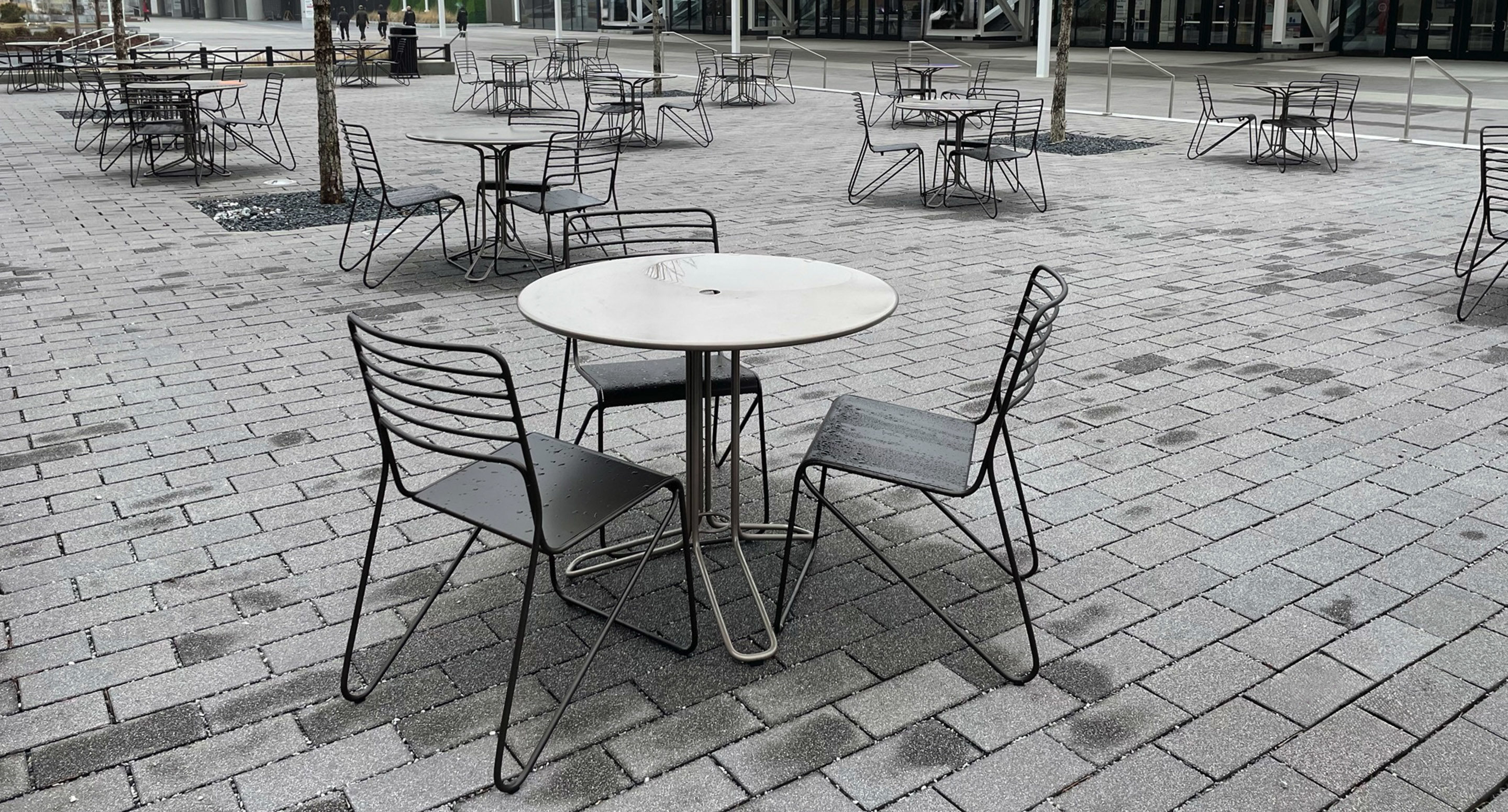 Empty metal tables and chairs are arranged on a paved outdoor plaza, with no people present and droplets of water visible on the furniture.