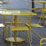 Several yellow metal tables and chairs are arranged on a paved outdoor patio area, with no people present.