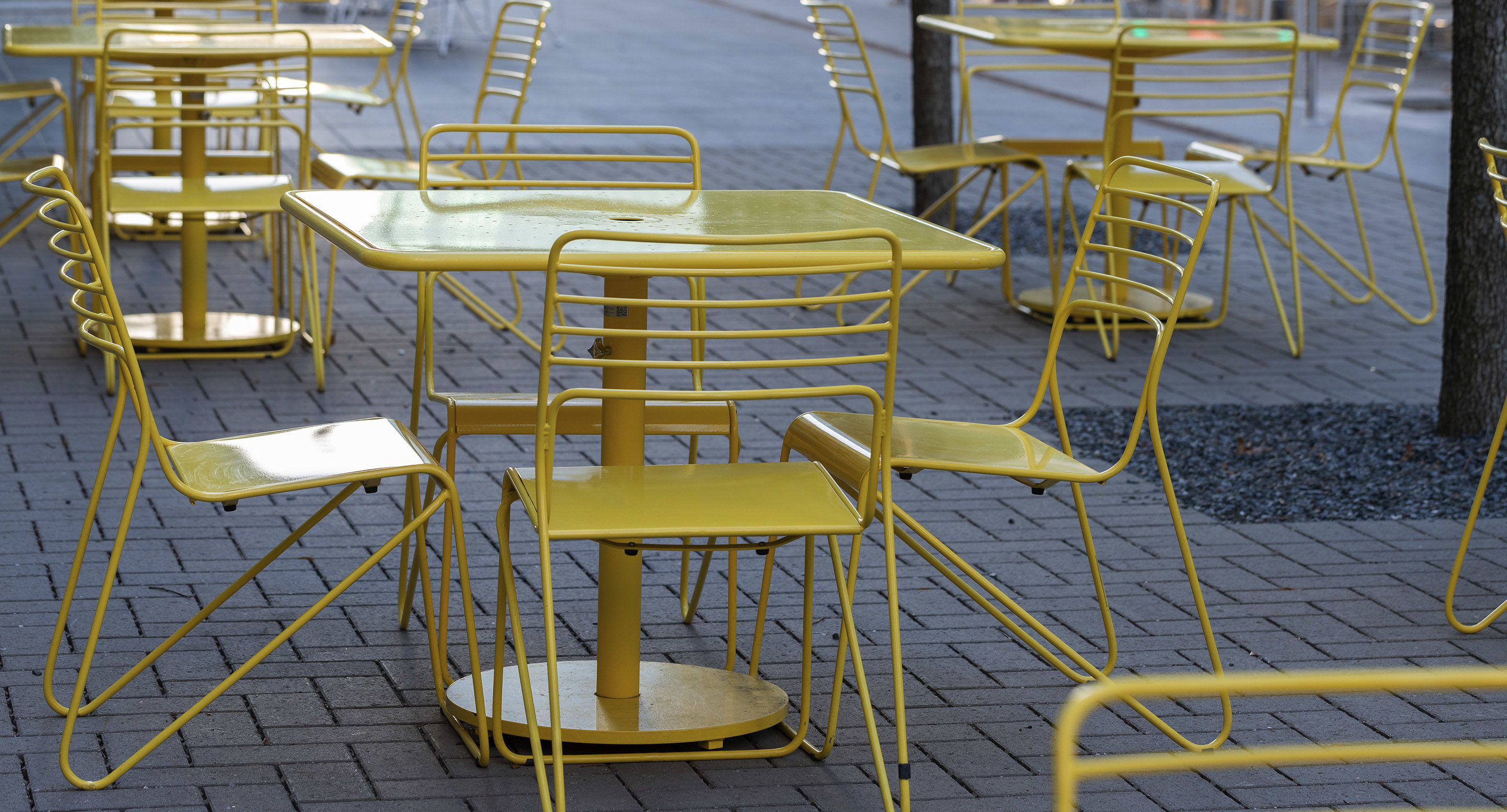 Several yellow metal tables and chairs are arranged on a paved outdoor patio area, with no people present.