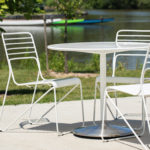 White metal table with three matching chairs on a concrete patio near a lake, with trees, grass, and a canoe in the background.