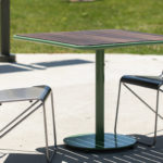 A small outdoor table with a wooden top and two metal chairs on a concrete surface, with grass visible in the background.