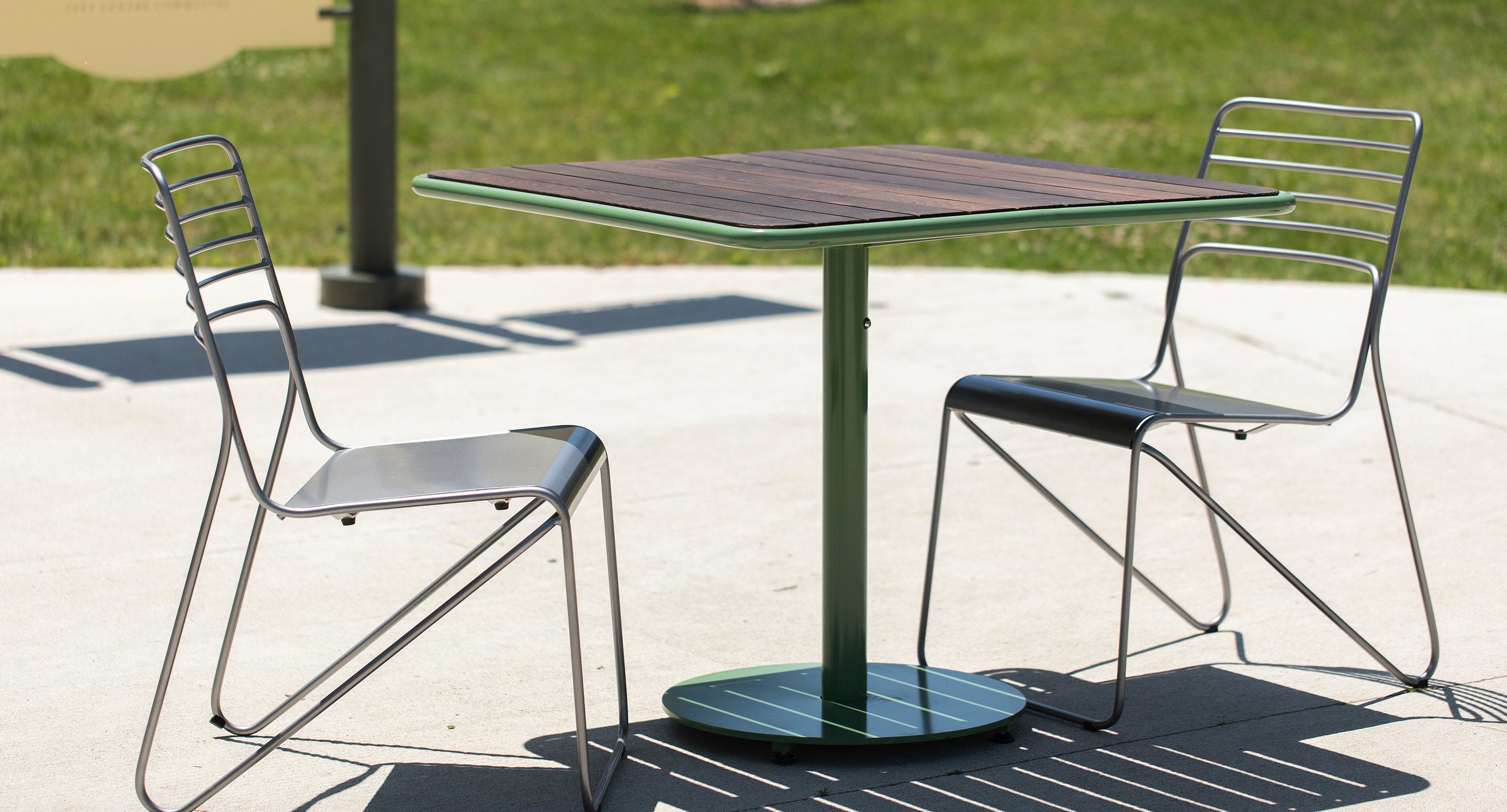 A small outdoor table with a wooden top and two metal chairs on a concrete surface, with grass visible in the background.