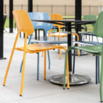 Four colorful plastic chairs and a black square table are arranged on a concrete patio under a covered outdoor area.