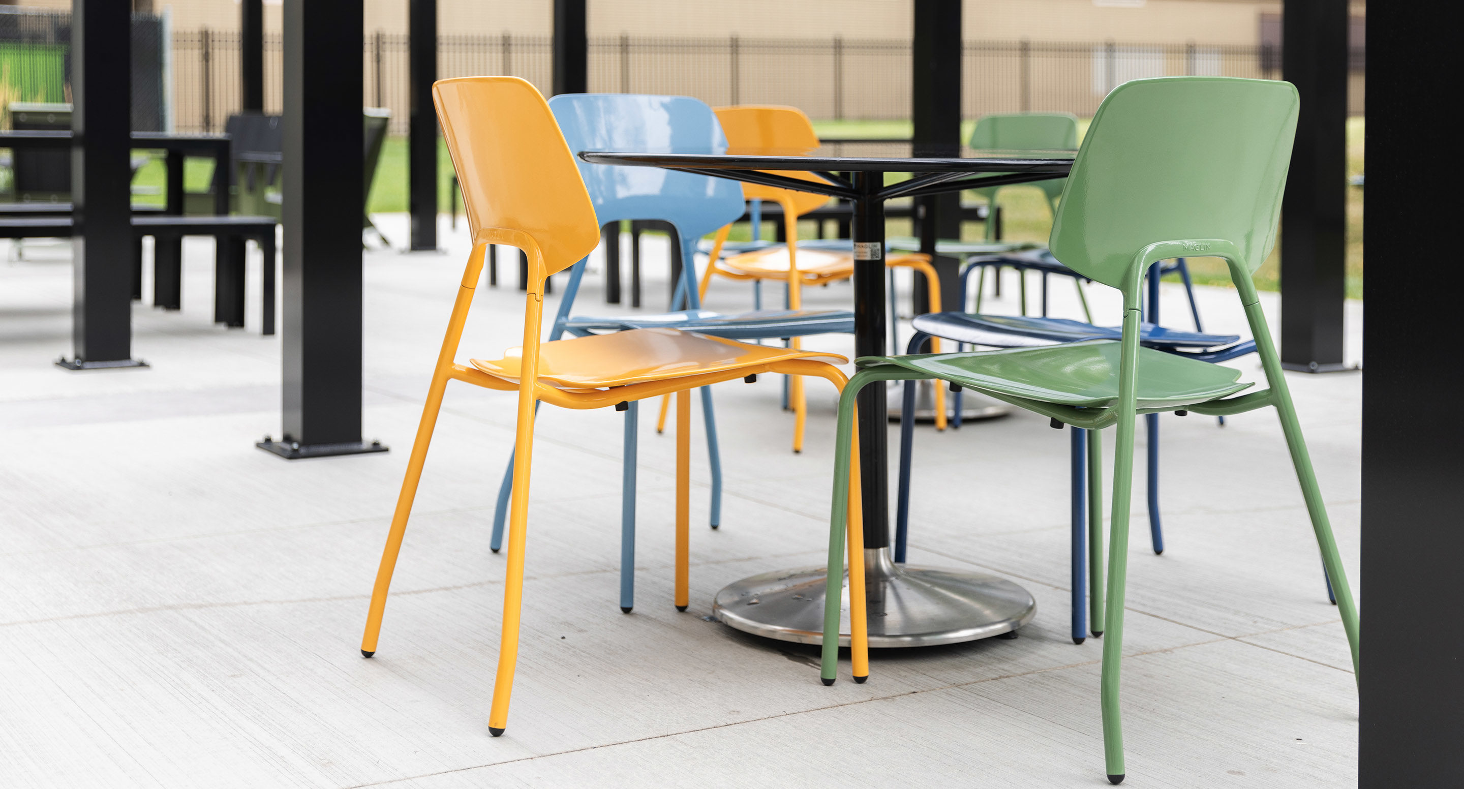 Four colorful plastic chairs and a black square table are arranged on a concrete patio under a covered outdoor area.