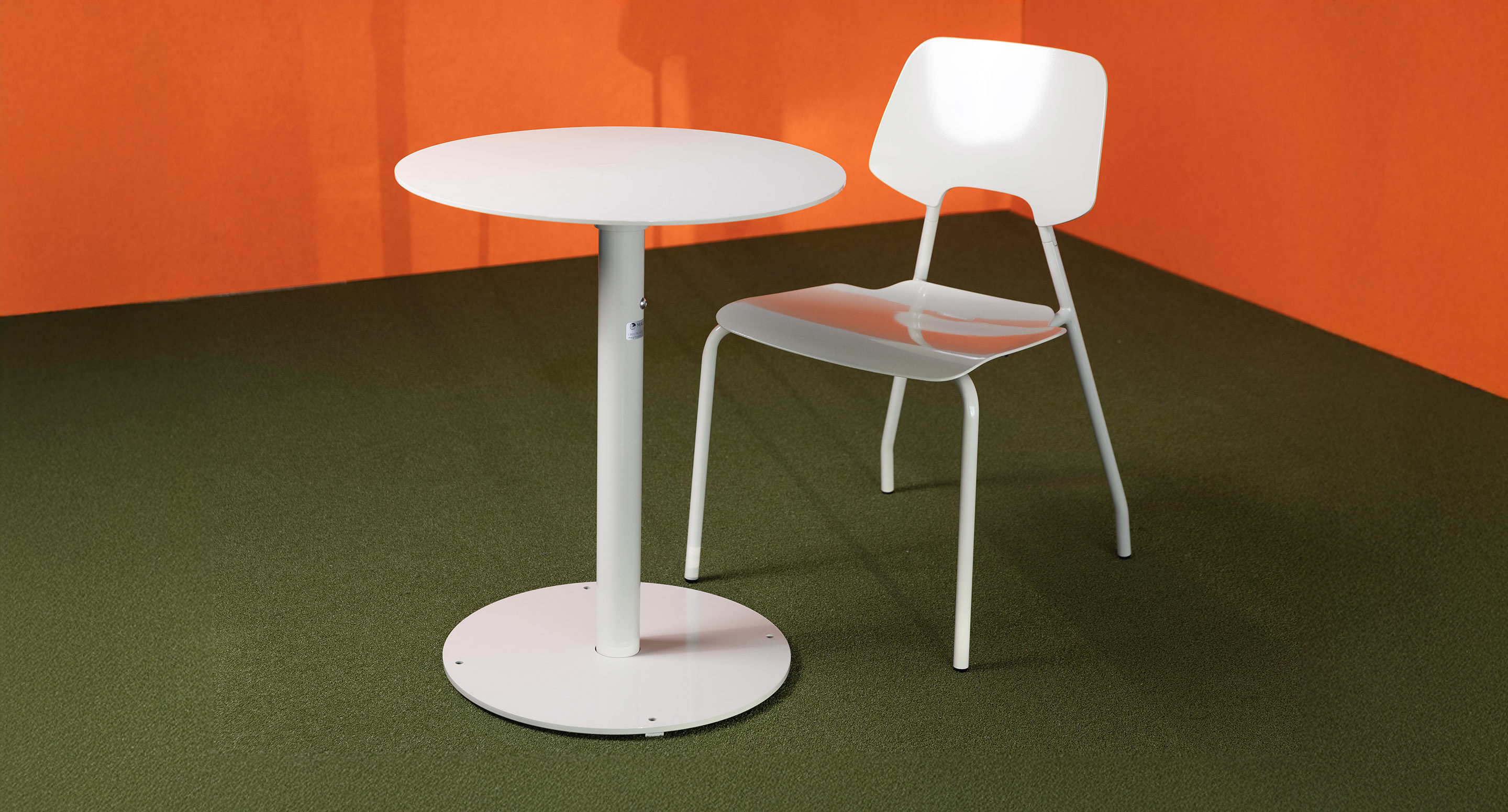 A white round table and a matching white chair are placed on a green carpet against an orange wall.