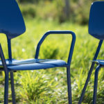 Two blue metal chairs with modern design are placed outdoors on grass, with sunlight illuminating the scene.