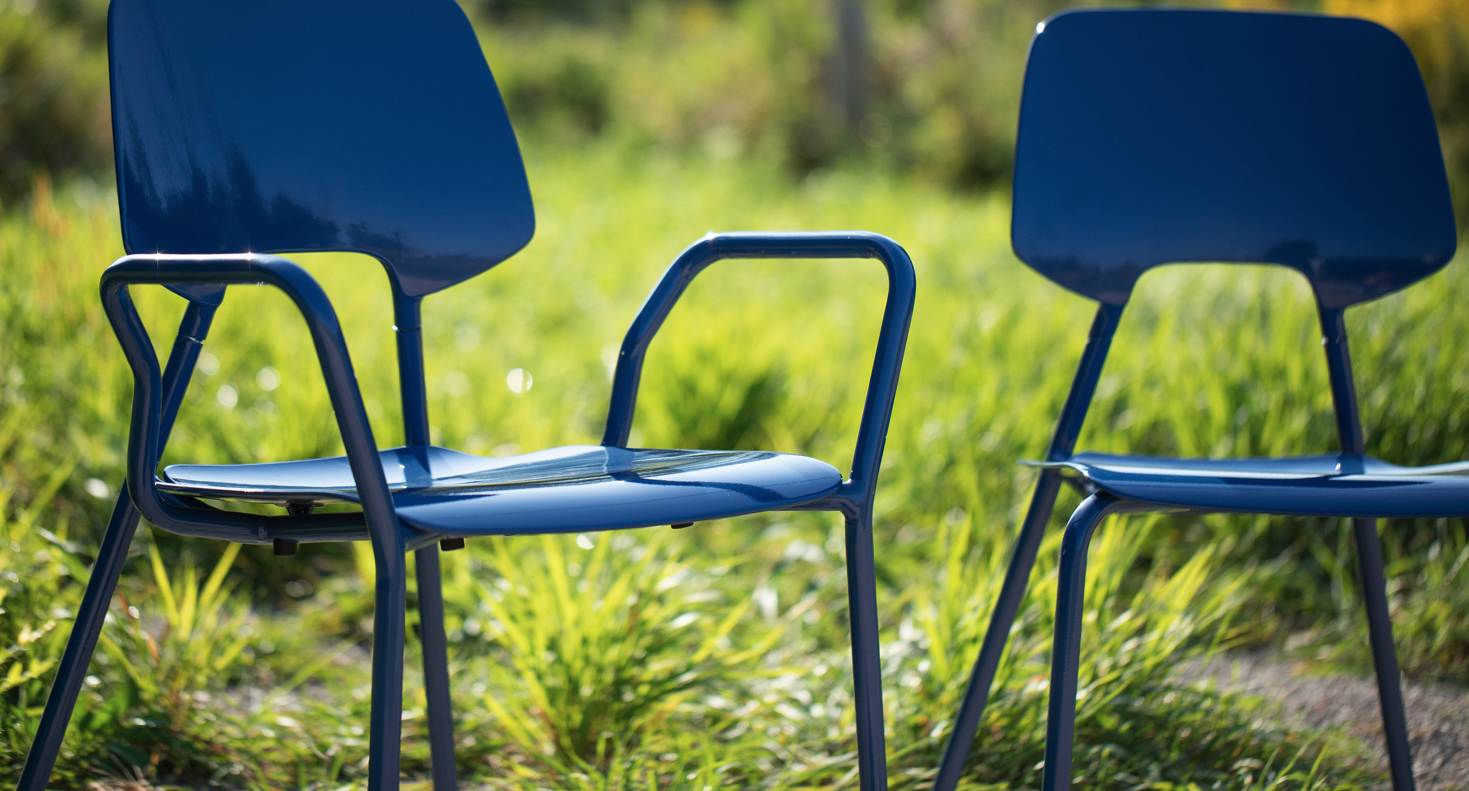 Two blue metal chairs with modern design are placed outdoors on grass, with sunlight illuminating the scene.