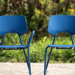 Two blue metal chairs are positioned outdoors on a gravel surface, with green foliage visible in the background.