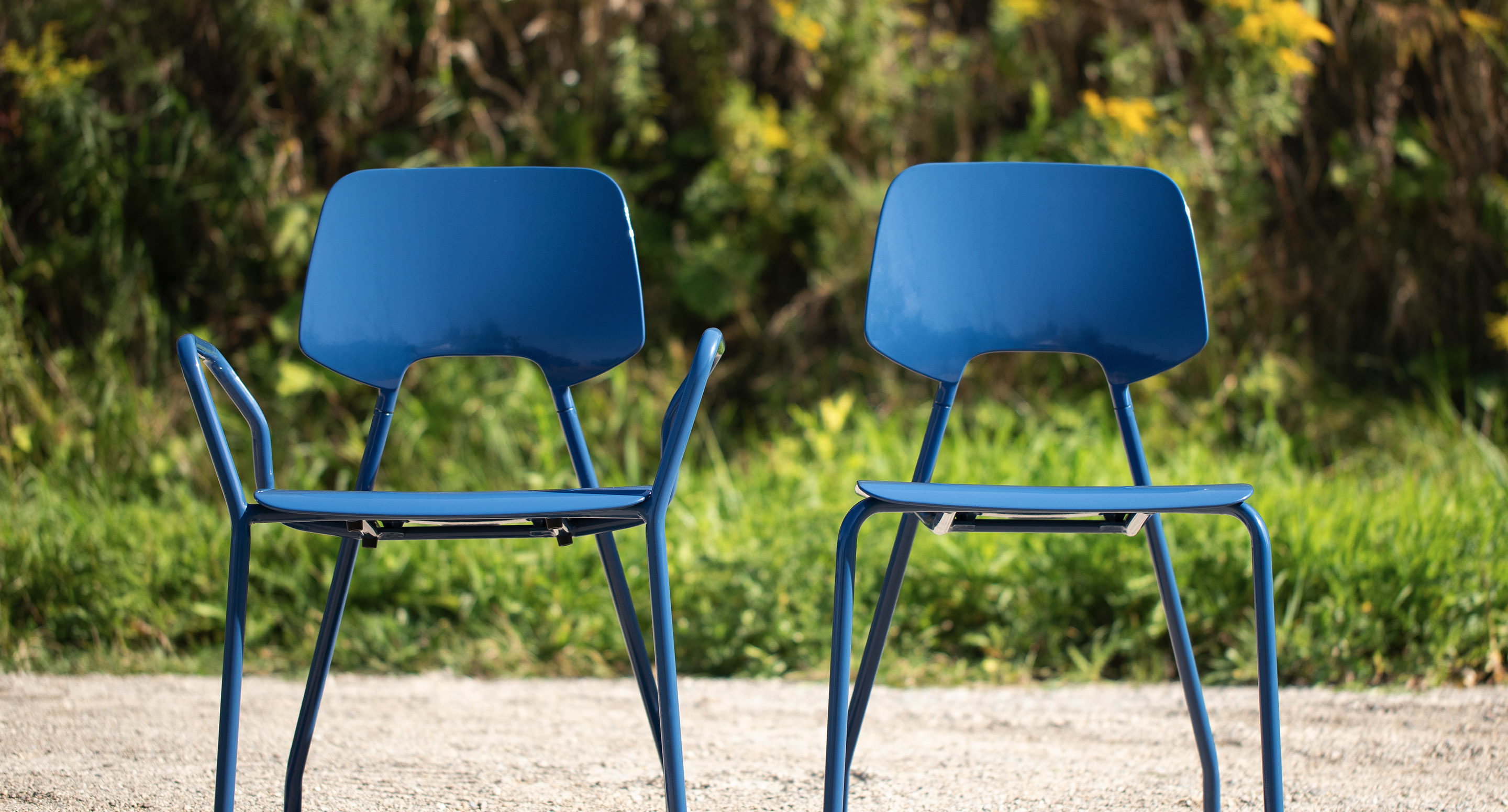 Two blue metal chairs are positioned outdoors on a gravel surface, with green foliage visible in the background.