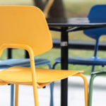 Four colorful chairs—yellow, green, and blue—surround a round black table outdoors, with grass and trees visible in the background.