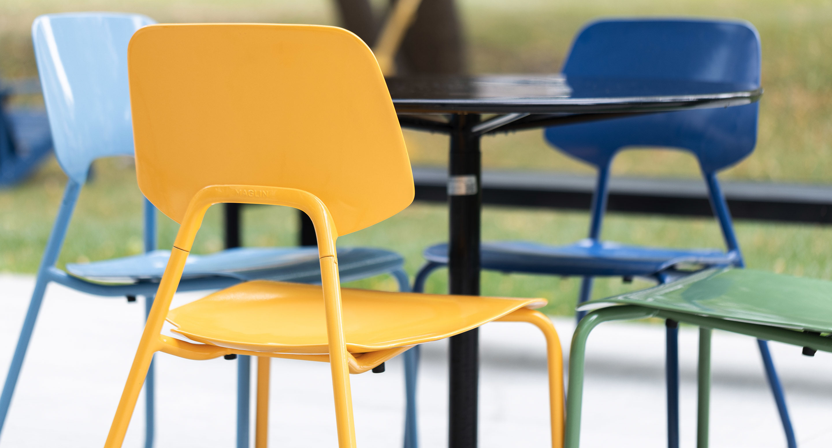 Four colorful chairs—yellow, green, and blue—surround a round black table outdoors, with grass and trees visible in the background.