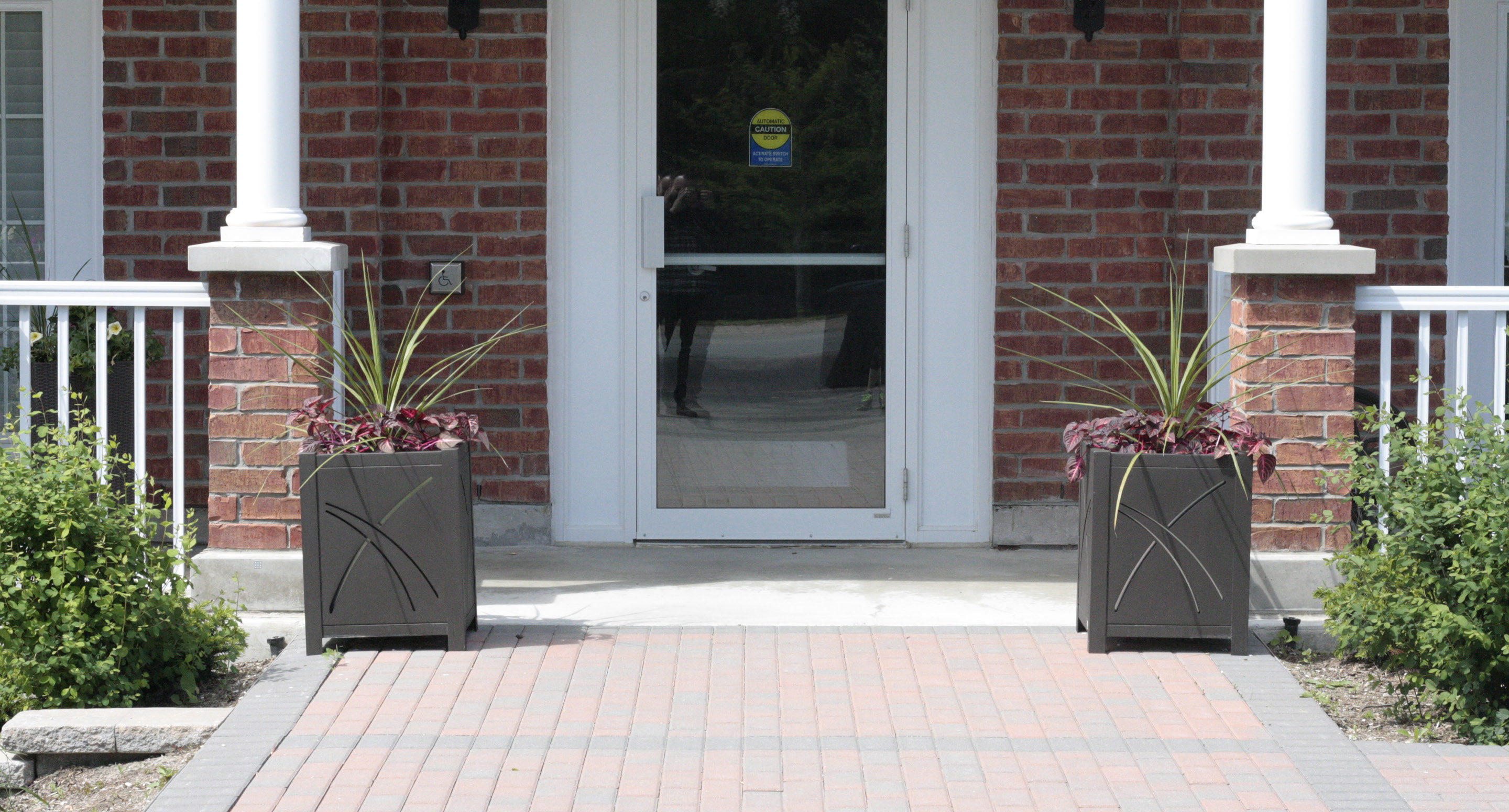 maglin_mpl-0400-planters-2 Une entrée principale avec une porte vitrée, des colonnes blanches, un mur de briques rouges, deux grandes jardinières carrées avec des plantes grimpantes et une allée pavée.