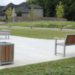 Un parc extérieur moderne avec un banc, une poubelle et des tables de pique-nique sur une surface en béton, entouré d'herbe, d'arbres et de maisons en arrière-plan.